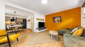 Uma sala de estar moderna com um sofá, uma mesa de centro e uma televisão. Dá para ver uma cozinha ao fundo, com armários e uma área de refeições.