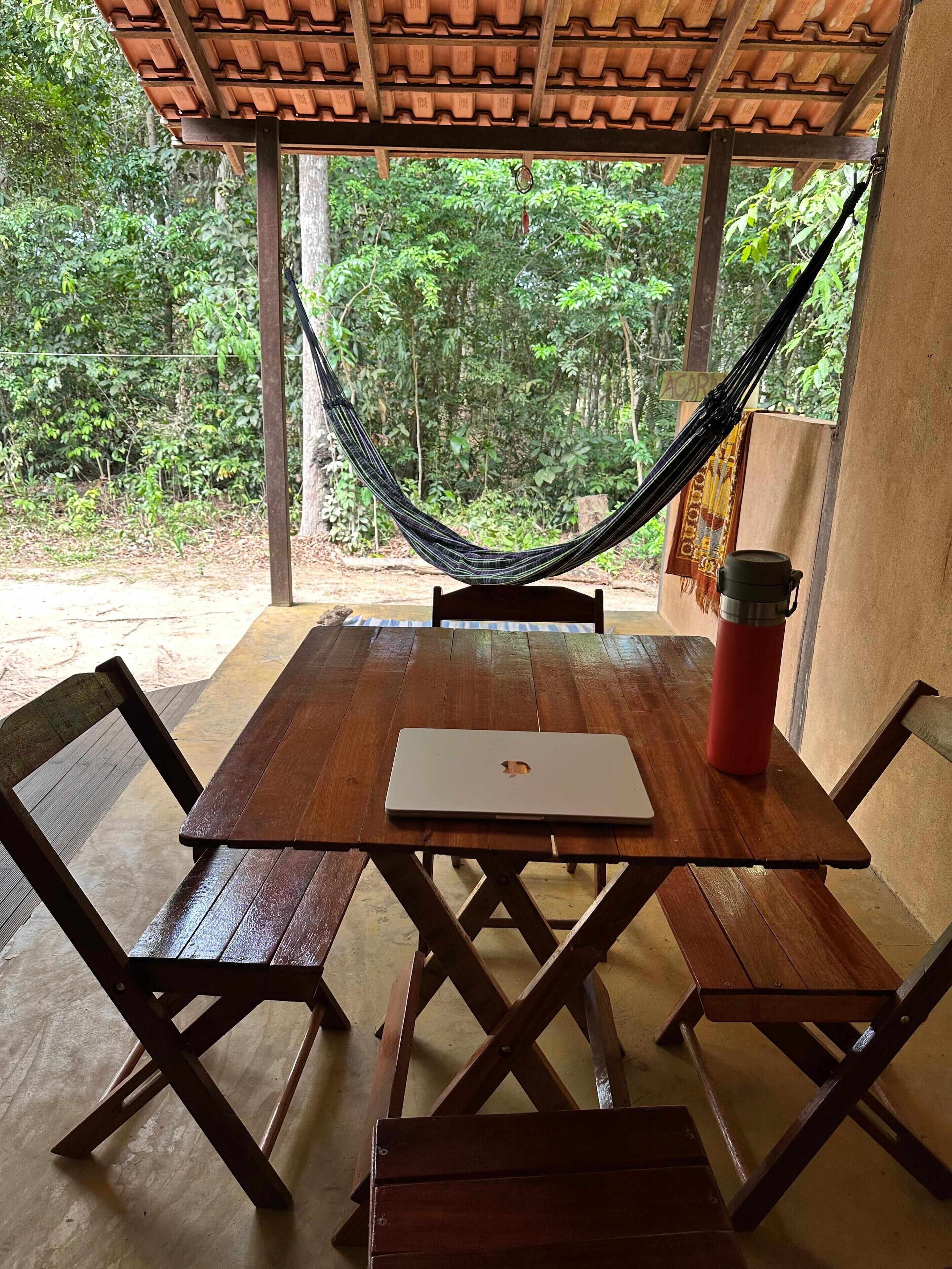 Une table en bois, sur laquelle se trouvent un ordinateur portable et un thermos rouge, placée sous une pergola en bois avec un hamac.