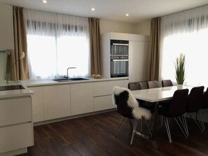 Eine moderne Küche mit Einbauofen, ein Essbereich mit weißem Tisch und Stühlen sowie große Fenster mit Vorhängen.