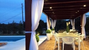 Un patio al aire libre con una mesa de madera y sillas blancas, rodeado de cortinas blancas y plantas en macetas.