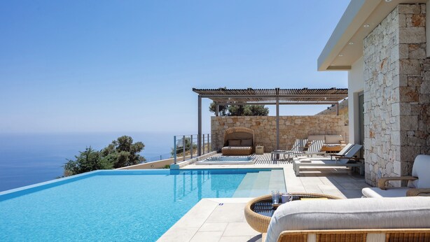 Una piscina infinita moderna con vista despejada al océano, mobiliario exterior con cojines y una pared de piedra.