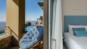 Un balcón con vistas a una piscina y al mar, una silla de mimbre y una cama con ropa blanca y almohadas azules.