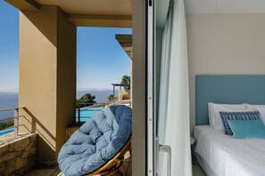 A balcony with a view of a pool and the sea, a wicker chair, and a bed with white bedding and blue pillows. - Melampes Villa, Seaview Escape, Heated Pool & Spa (Rethymno)