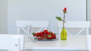 Une table blanche ornée d’un tableau représentant un paysage côtier, un verre de vin, une assiette de fraises et un vase contenant une seule rose rouge.