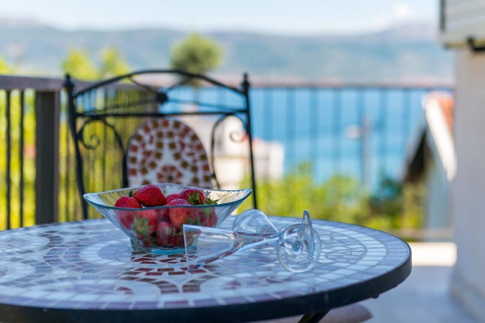 Un saladier en verre transparent contenant des fraises, posé sur une table à motifs de mosaïque, avec une chaise en métal, et en arrière-plan, une vue sur des montagnes et des bâtiments.