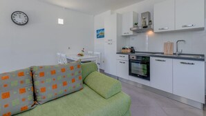 Une cuisine moderne avec des meubles blancs, un four encastré et un évier. Le salon est agrémenté d’un canapé vert avec des coussins aux motifs orange et verts.