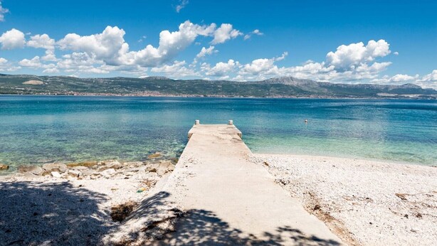 Un quai en béton s’étendant dans une mer d’un bleu limpide, avec une côte rocheuse et des collines lointaines.