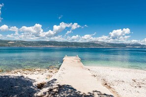 Ein Betonsteg erstreckt sich in ein klares, blaues Meer mit einer felsigen Küste und entfernten Hügeln.
