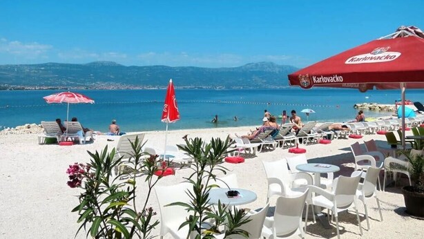 A beachside restaurant with outdoor seating, a view of the sea, and a red umbrella branded with 'Karolivacko'. - Two Bedroom Apartment with Shared Pool (Arbanija)