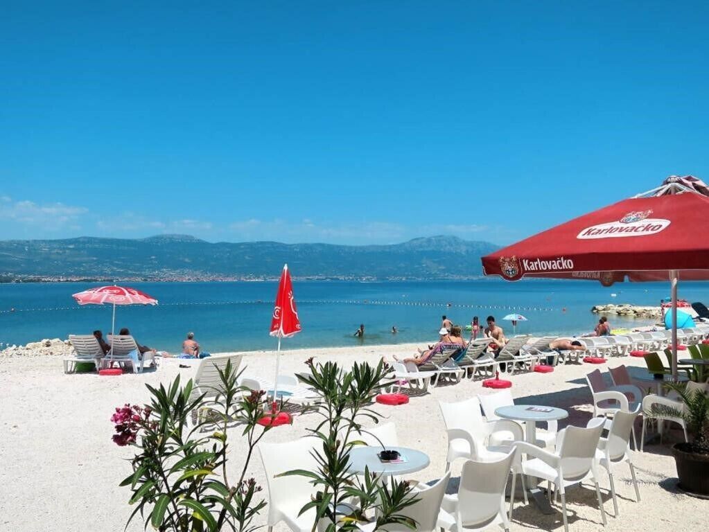 A beachside restaurant with outdoor seating, a view of the sea, and a red umbrella branded with 'Karolivacko'.
