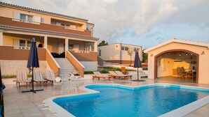 Une piscine avec des chaises longues et des parasols, un escalier menant à un balcon, et un bâtiment doté d’un toit plat et de murs de couleur beige.