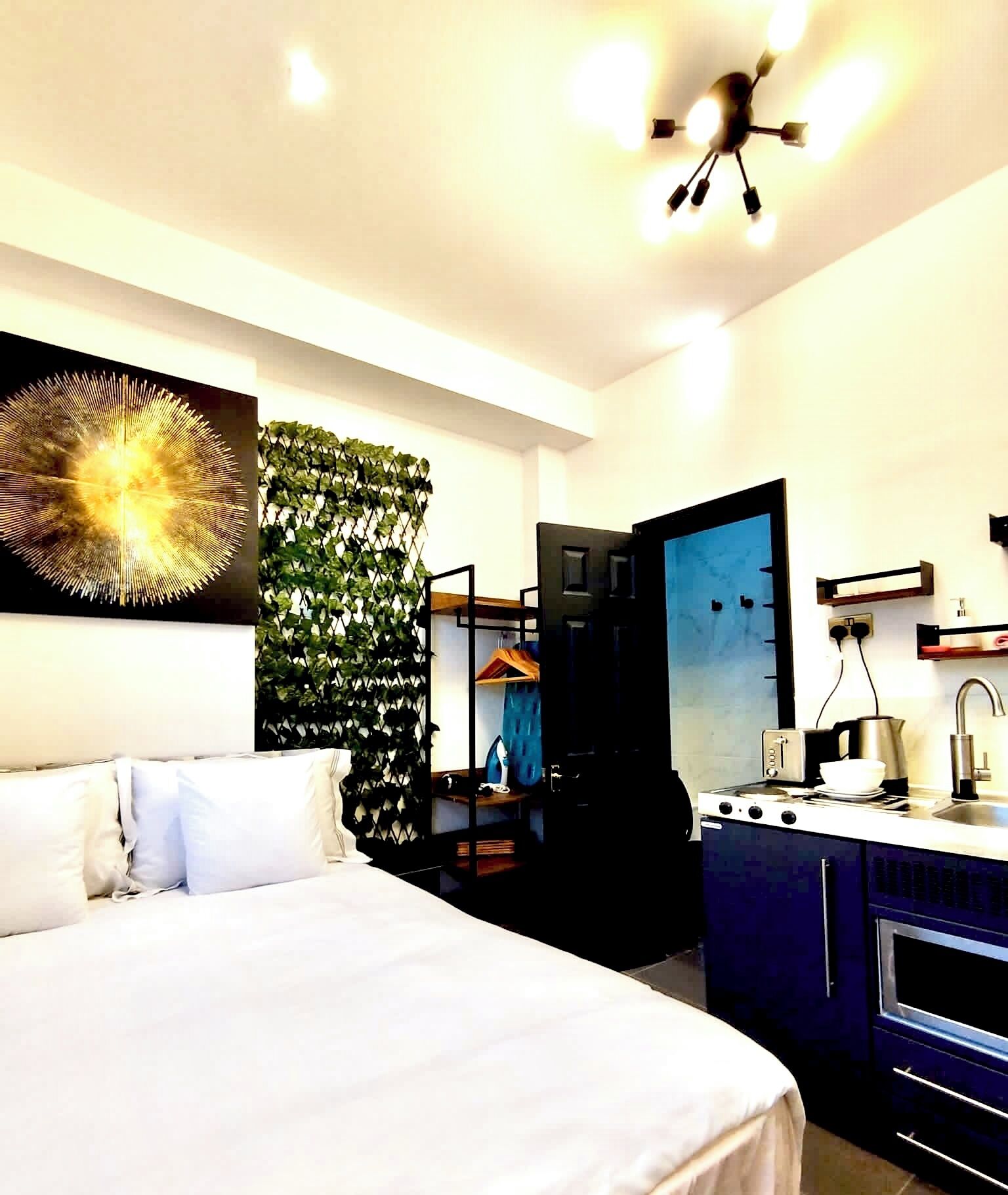 A modern hotel room with a bed, a kitchenette, a mirror, and a wall-mounted artwork.
