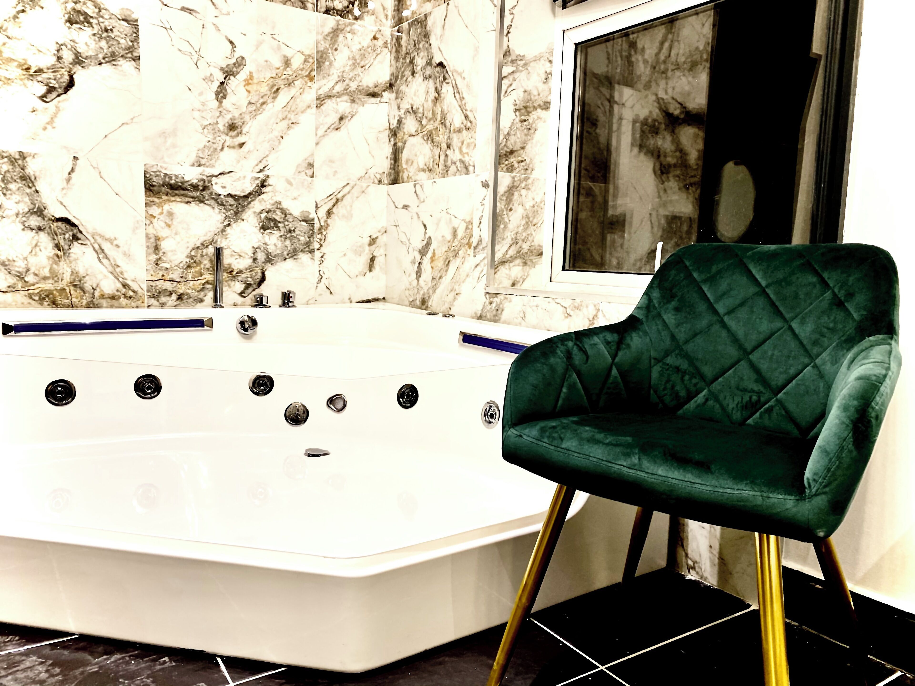 A bathroom with a marble wall, a white bathtub, and a green chair with gold legs.