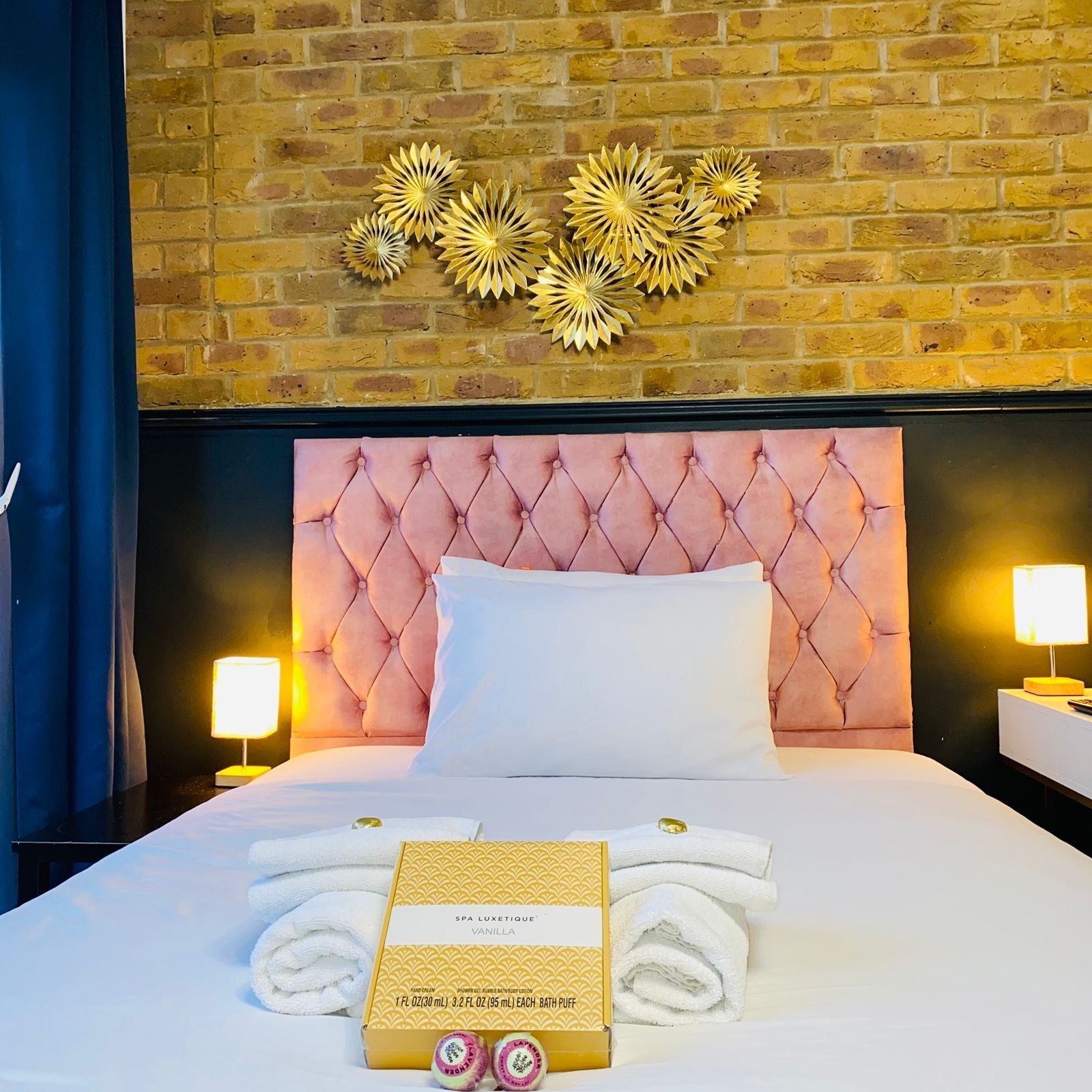A hotel room with a brick wall, a tufted headboard, two bedside lamps, a neatly made bed with white linens, and a box of spa towels.