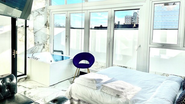 A modern bedroom with a large bed, a bathroom with a bathtub, and a city view through large windows. - BPR Private Hot-tubs and rooms  (London)