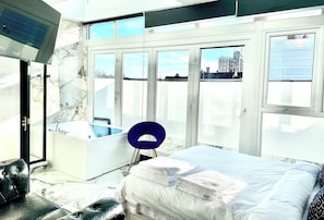 A modern bedroom with a large bed, a bathroom with a bathtub, and a city view through large windows. - BPR Private Hot-tubs and rooms  (London)