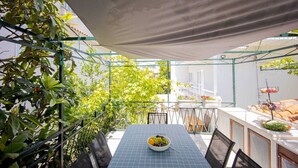 Un espace extérieur abrité, aménagé pour les repas, avec une table pour deux, entouré de verdure et bordé d’un mur blanc.
