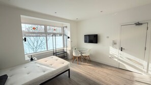 A modern bedroom with a bed, a TV, a small table with chairs, and a window with a view of the outside. - Modern Studio Flat in Streatham | Close to Central London (London)