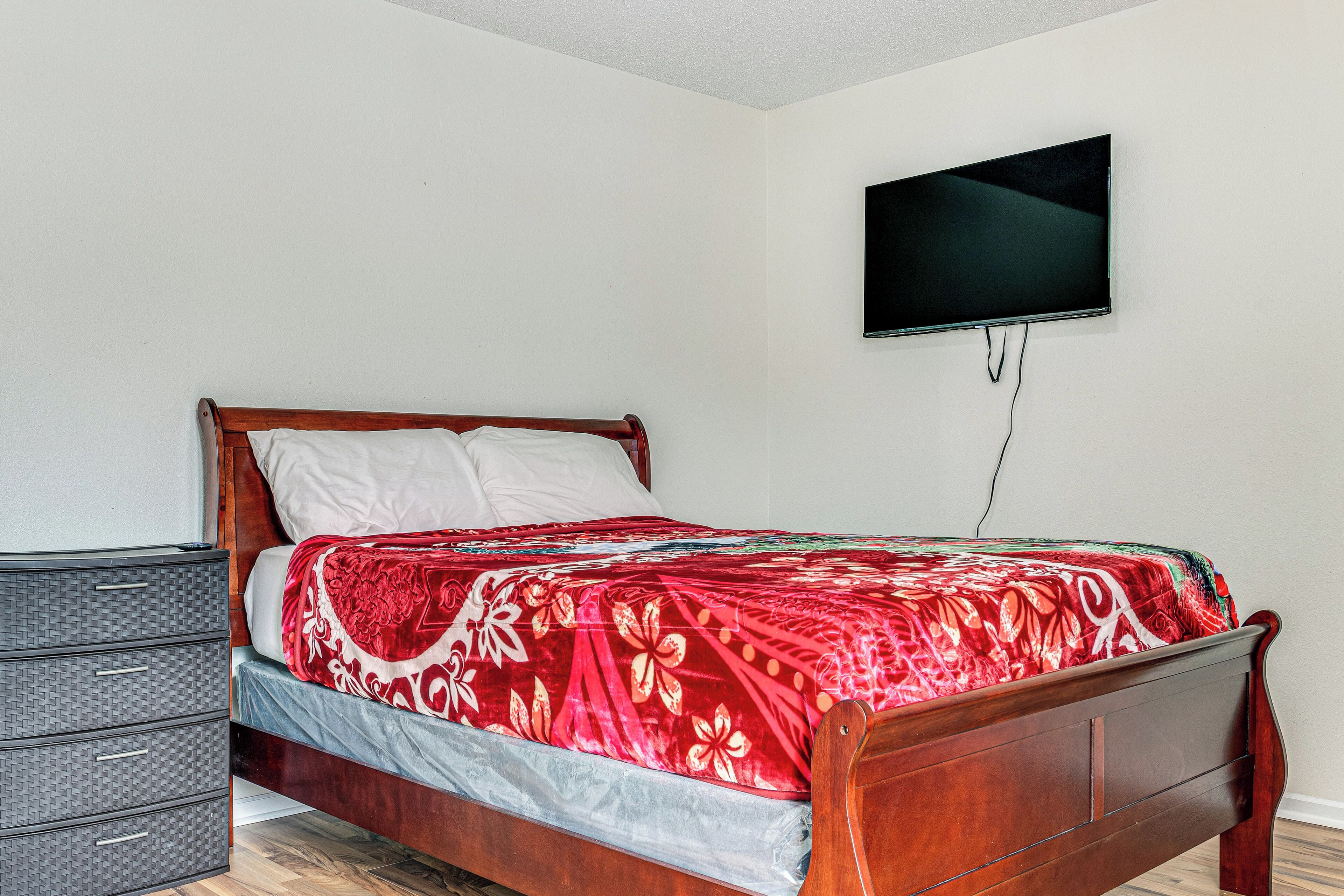 A neatly made bed with a red floral bedspread, a wooden headboard, and a television mounted on the wall.