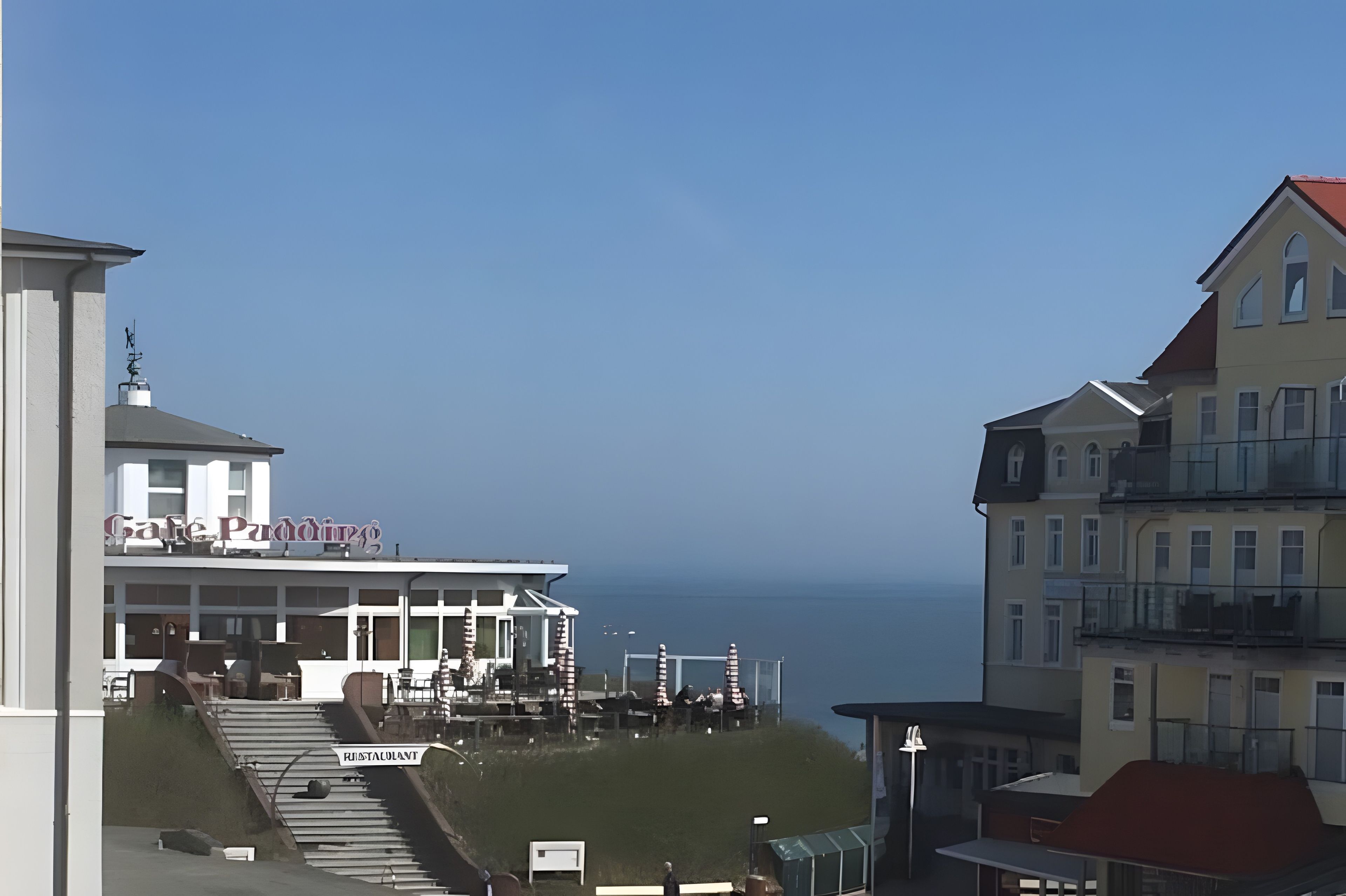 A coastal building with a balcony overlooking the sea, a sign indicating a restaurant, and a clear sky.