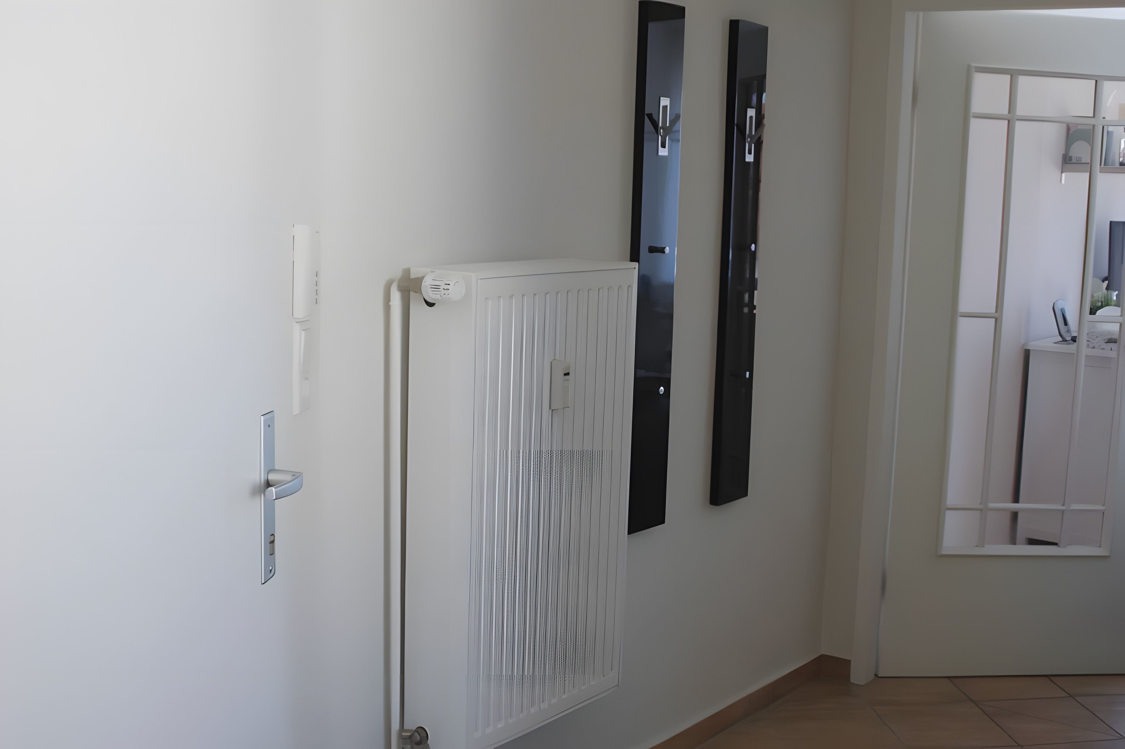 Ein Flur mit einem weißen Heizkörper, einer Tür mit moderner Griffleiste und zwei vertikalen schwarzen Objekten an der Wand.