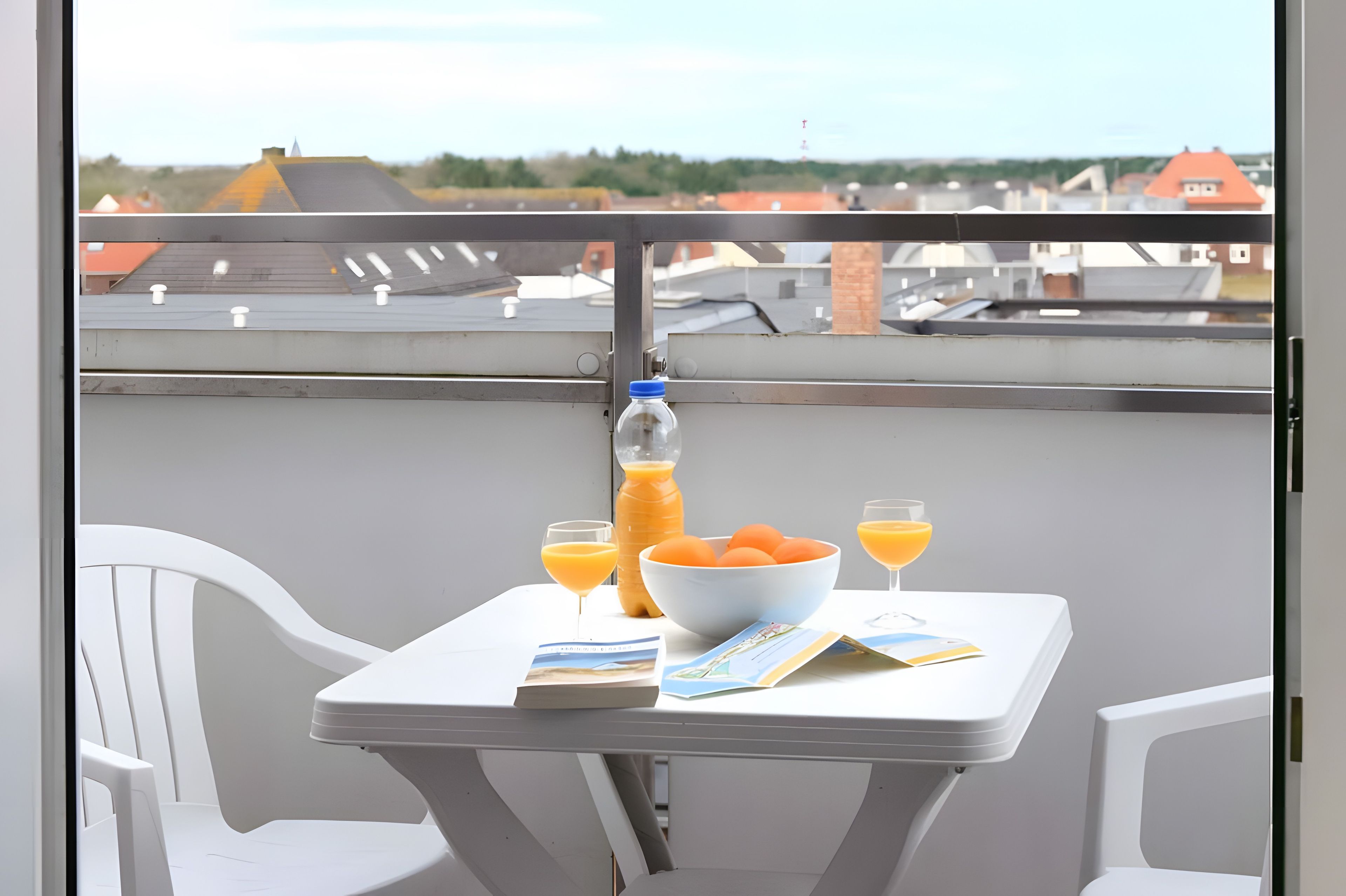 A balcony with a table set for breakfast, including a bottle, a bowl of fruit, and glasses of orange juice.
