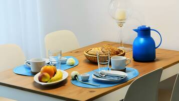 A breakfast table with a fruit bowl, a glass of water, a cup, a jug, and a basket of bread.