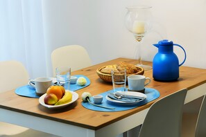 Una mesa de desayuno con un bol de fruta, un vaso de agua, una taza, una jarra y una cesta de pan.