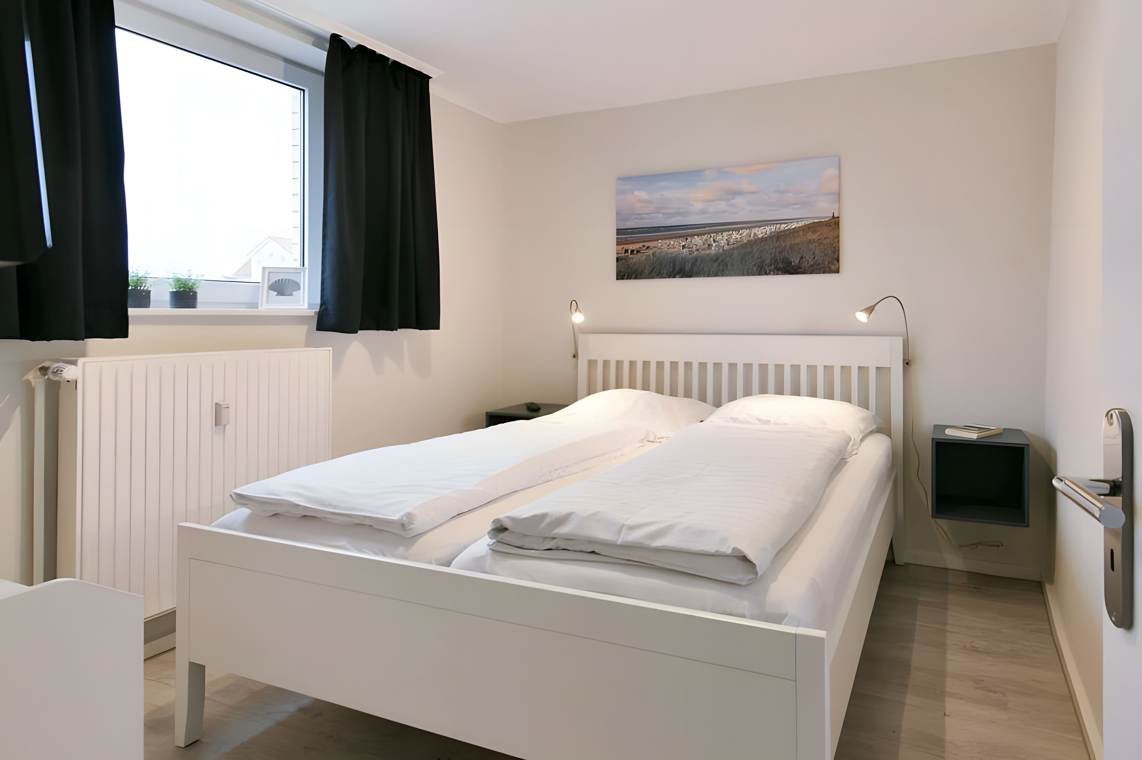 A neatly made twin bed with white linens, a bedside table with a lamp, a wall-mounted TV, and a window with black curtains.
