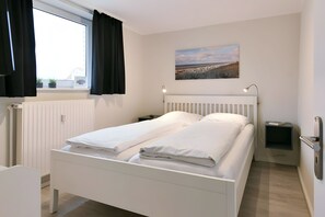 Un lit jumeau bien fait, avec du linge de lit blanc, une table de chevet avec une lampe, un téléviseur fixé au mur et une fenêtre avec des rideaux noirs.