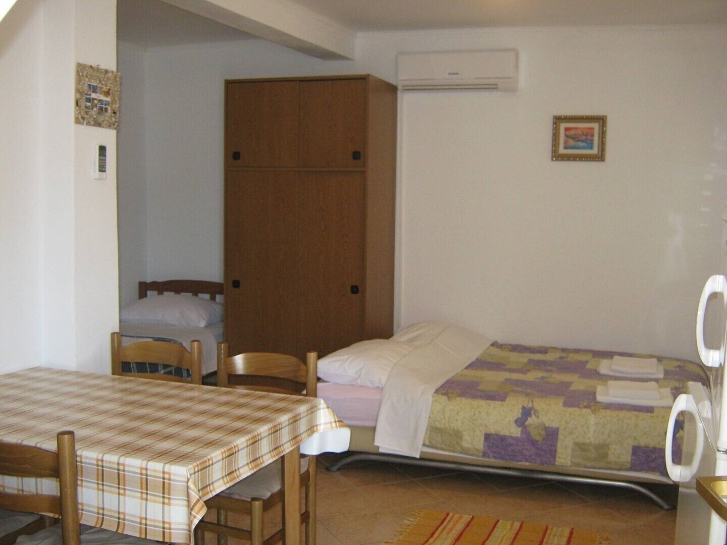 A small room with a bed, a table, chairs, a wardrobe, and an air conditioning unit.