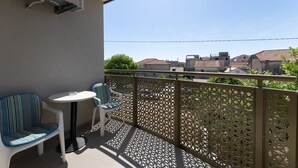 Un balcón con una mesa blanca y dos sillas a rayas, una valla ornamental de metal y vistas de edificios al fondo.