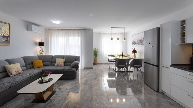 Una sala moderna con un sofá gris, una mesa de centro y un comedor con una mesa y sillas. Se observa una cocina con un refrigerador y armarios.