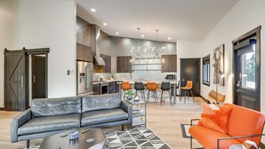 Un salon moderne avec un canapé gris, un fauteuil orange et un tapis à motifs géométriques. On aperçoit, au fond, une cuisine dotée d’un pare-feu hexagonal et d’appareils électroménagers en acier inoxydable.