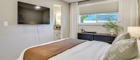 A bedroom with a bed, a TV mounted on the wall, a window with curtains, and a nightstand with a lamp.
