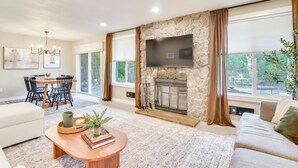 A living room with a stone fireplace, a flat-screen TV, a dining area with a table and chairs, and a large window with curtains. - Peace & Serenity in Gresham! Sleek Home w/ Yard (Gresham)