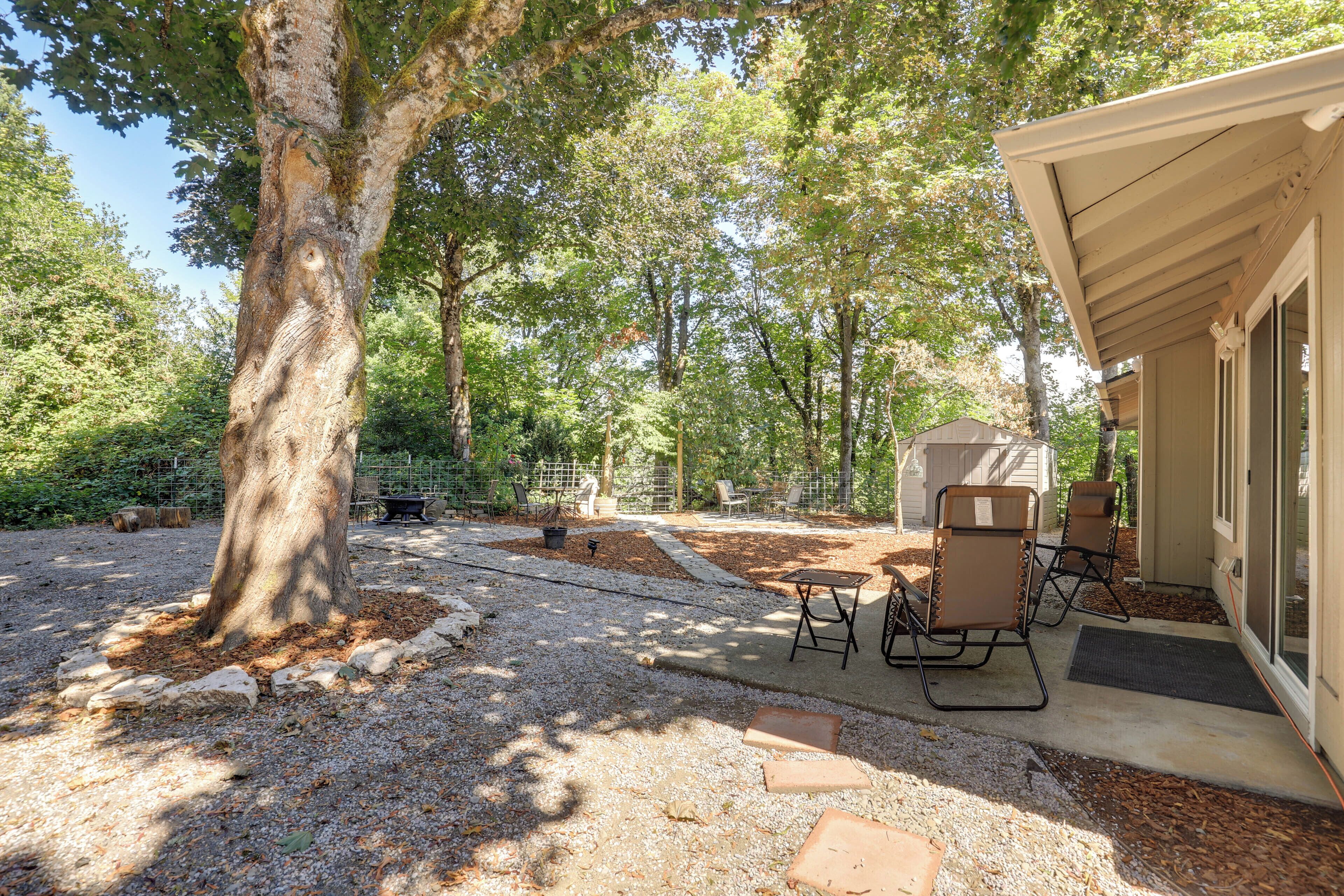 A covered outdoor seating area with a table and chairs, surrounded by trees and a gravel path.