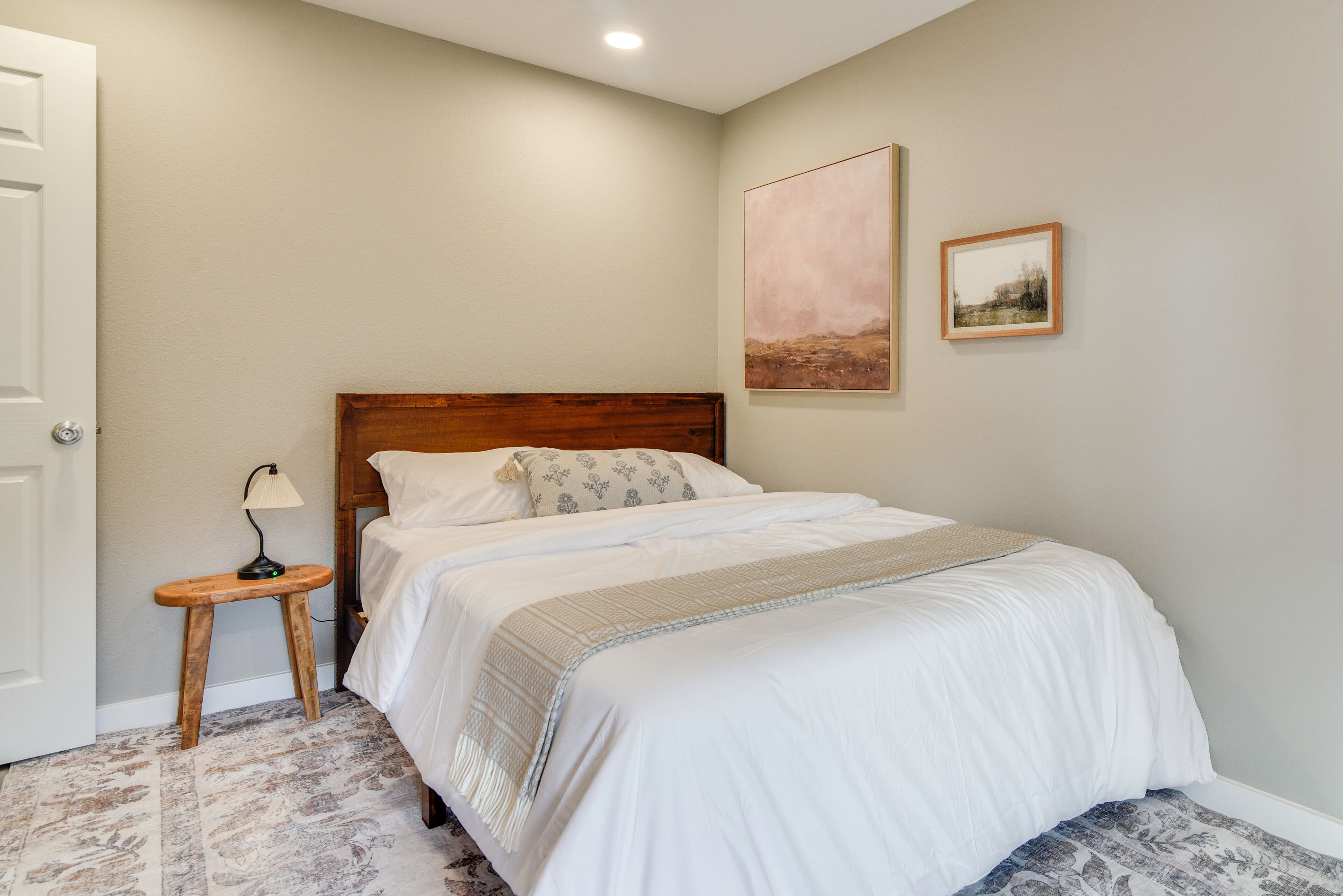 A bedroom with a wooden headboard, a bed with white bedding, a small bedside table with a lamp, and two framed pictures on the wall.