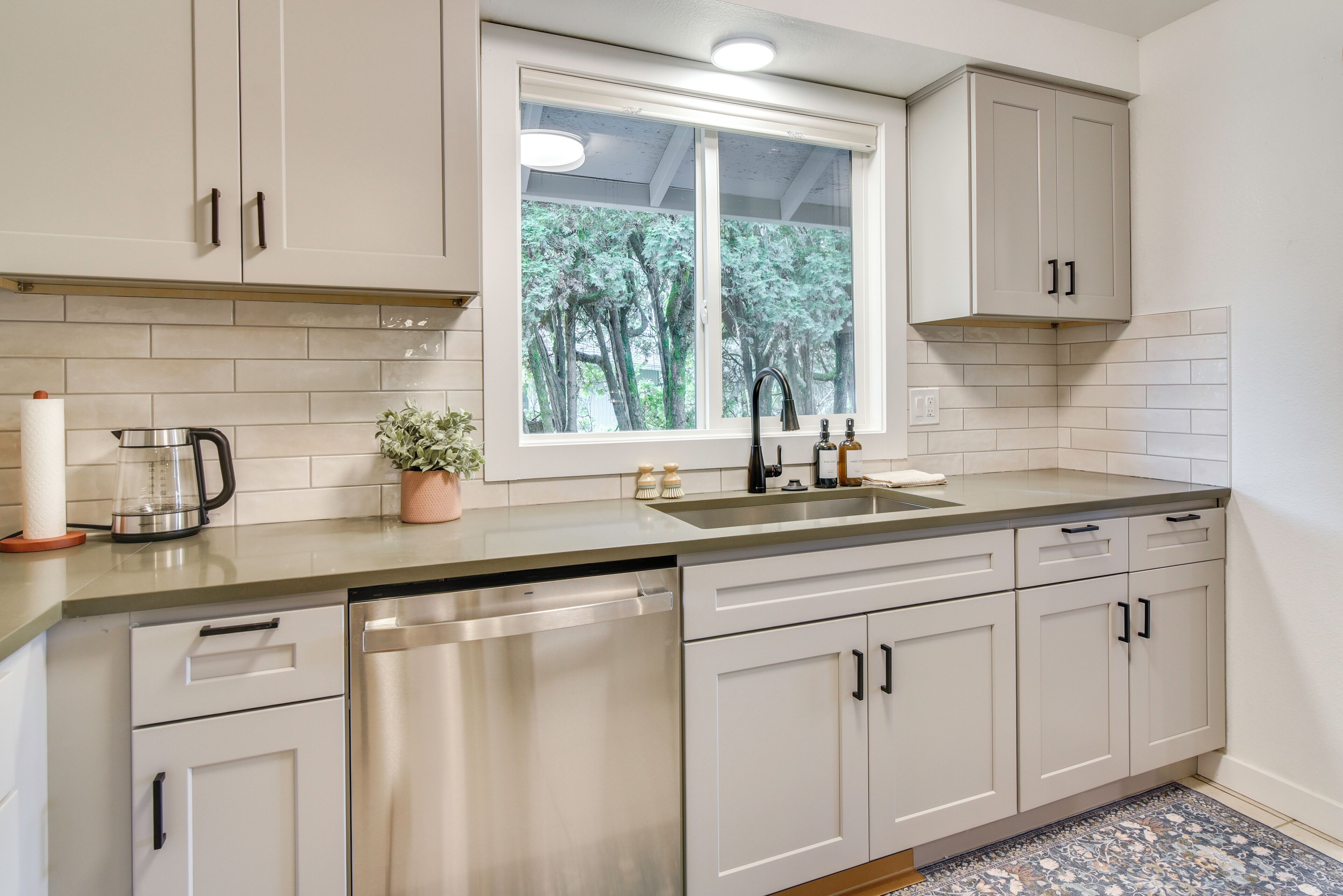 A kitchen with white cabinets, a stainless steel dishwasher, a window with a view of trees, and a countertop with a kettle and a plant.