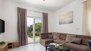 A modern living room with a brown sofa, a coffee table, and a TV. There is a sliding glass door leading to a balcony with curtains. - Two Bedroom Apartment (Kaštel Sućurac)