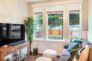 Ett vardagsrum med en tv-apparat på en träkonsol, ett stort fönster med persienner, en grå soffa med mönstrade kuddar och en krukväxt.
