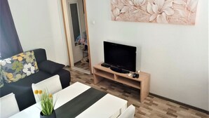 Ein Wohnzimmer mit einer Couch, einem Fernseher auf einem Holzschrank, einem Esstisch mit einer Pflanze und einem Blumenbild an der Wand.