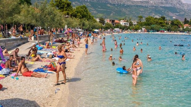 Ein voller Strand mit Menschen, die sich sonnen und schwimmen, umgeben von Bergen und Gebäuden.