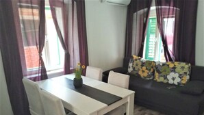 Ein Essbereich mit weißem Tisch und Stühlen, einer Couch mit floralen Kissen und einem Fenster mit violetten Vorhängen.