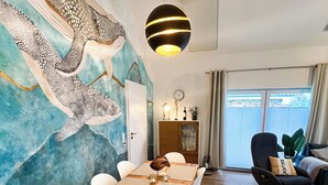 Un espace repas comprenant une table en bois, des chaises blanches, une fresque murale et un luminaire suspendu moderne.