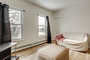 Quarto com duas janelas, porta branca, cama com cobertura branca, uma mesinha e um banco.