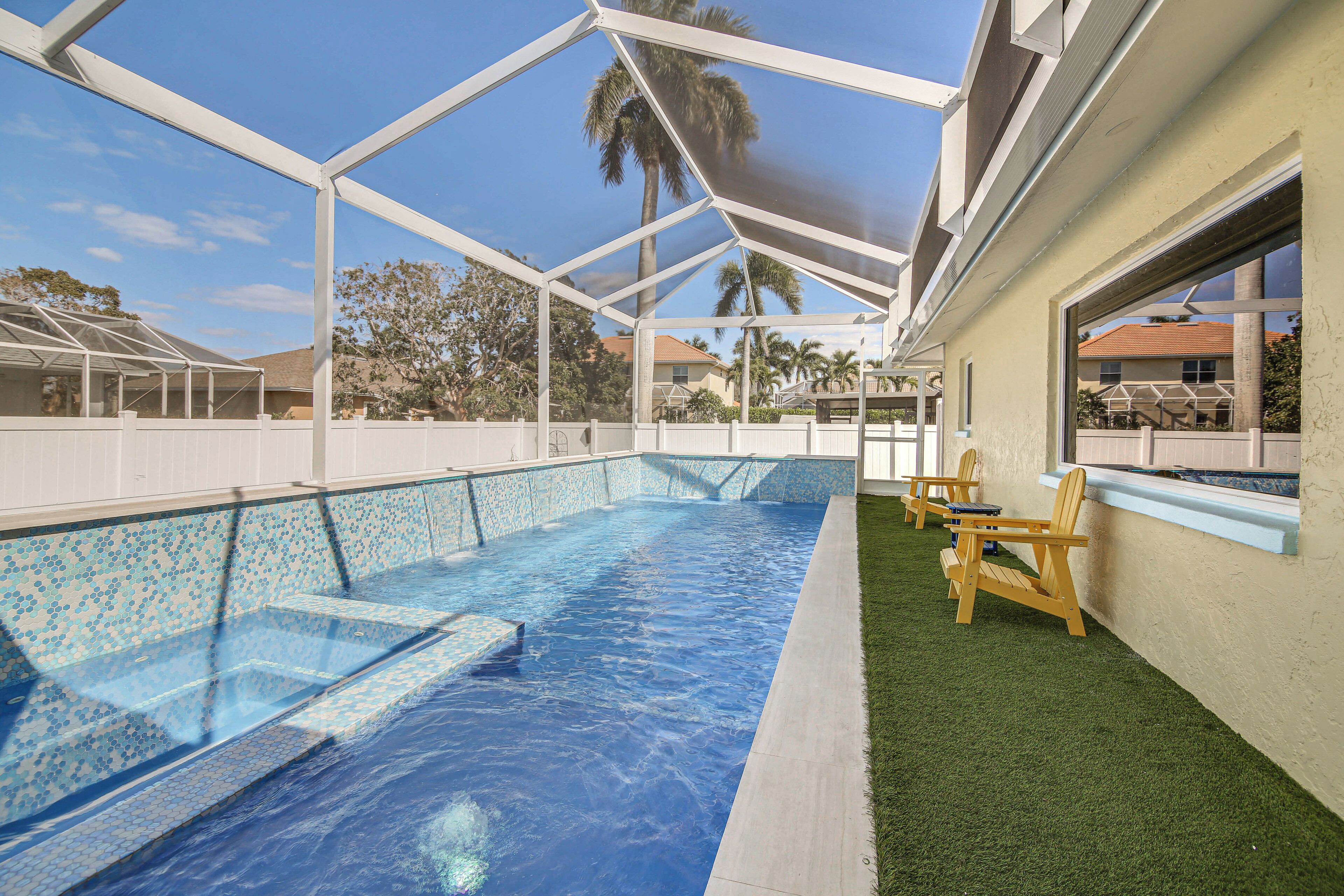 Una piscina con cerramiento de vidrio, rodeada de césped artificial y sillas de madera.