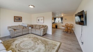 A living room with a sectional sofa, a dining area with a table and chairs, a kitchen in the background, and a mounted TV on the wall.