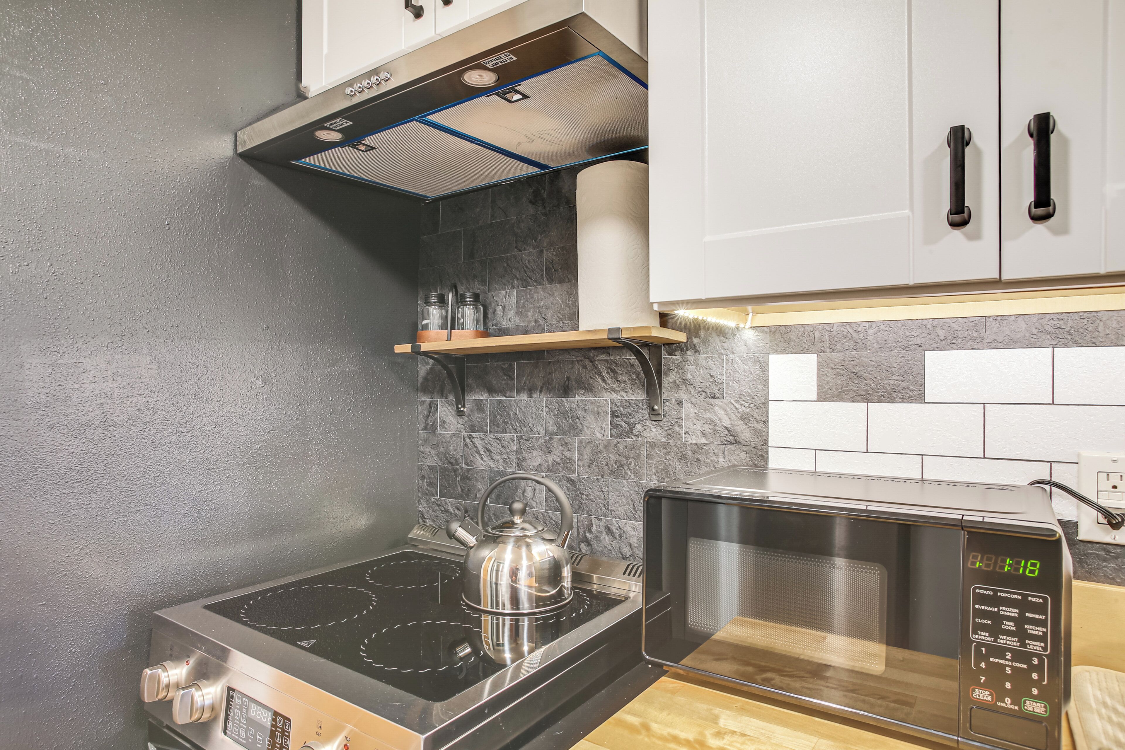 A kitchen with a stove, kettle, microwave, and cabinets.