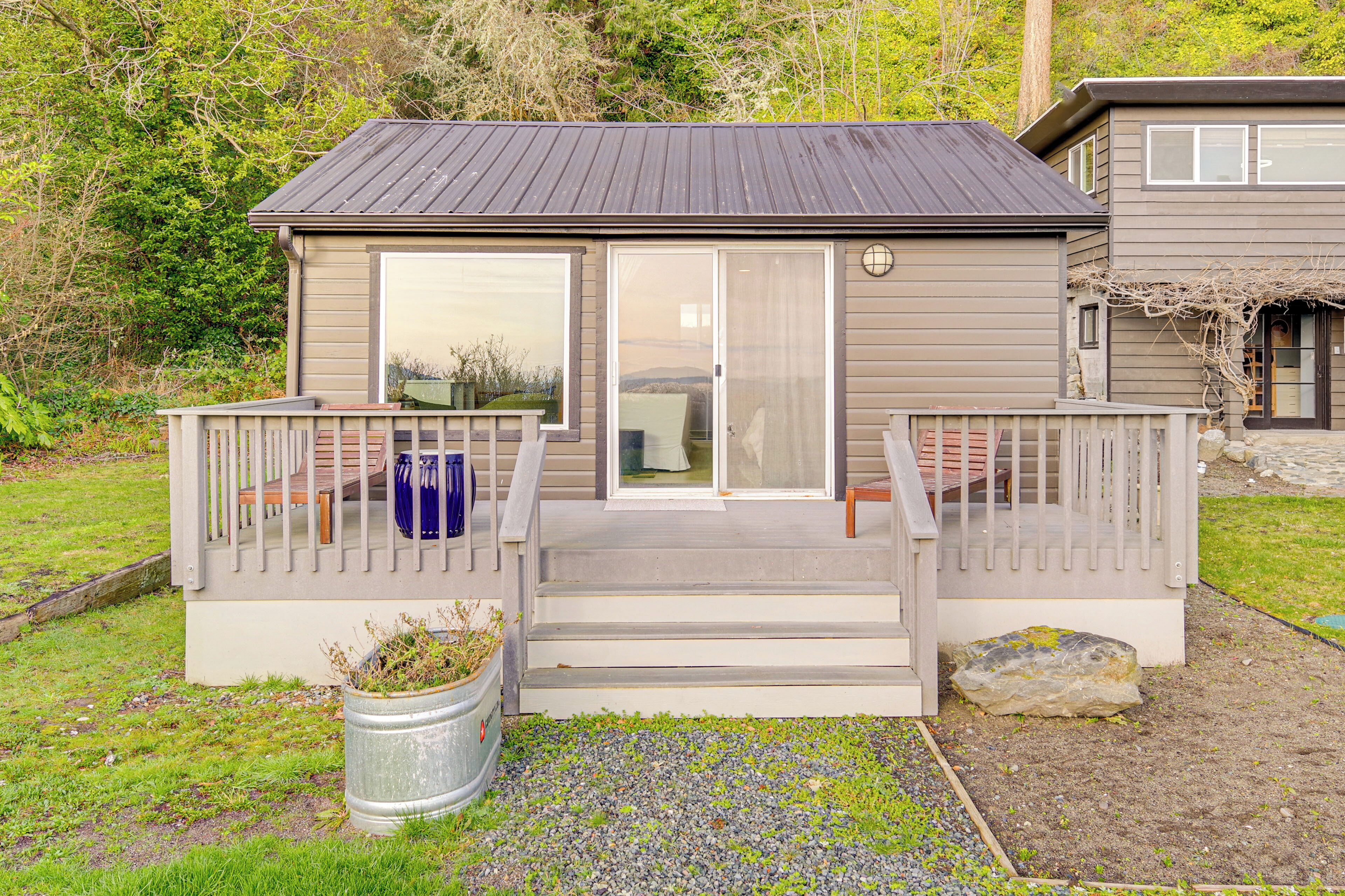 A small, single-story house with a deck, large sliding glass doors, and a stone pathway leading to the entrance.